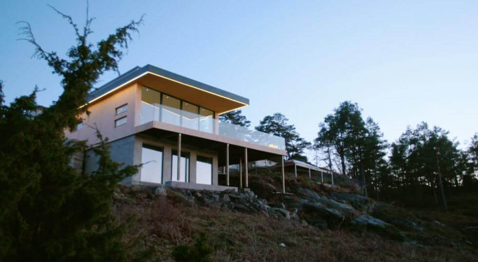 Stiligt - Stiligt modernt hus med stora fönster och platt tak, beläget på en klippig sluttning omgiven av träd under klar himmel.