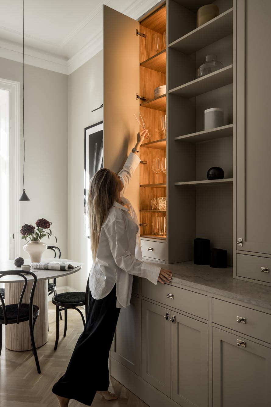 Stiligt - En person i stiligt vit skjorta sträcker sig efter glas i ett högt köksskåp. Rummet har ett matbord med stolar och en vas med blommor som utstrålar en elegant charm.